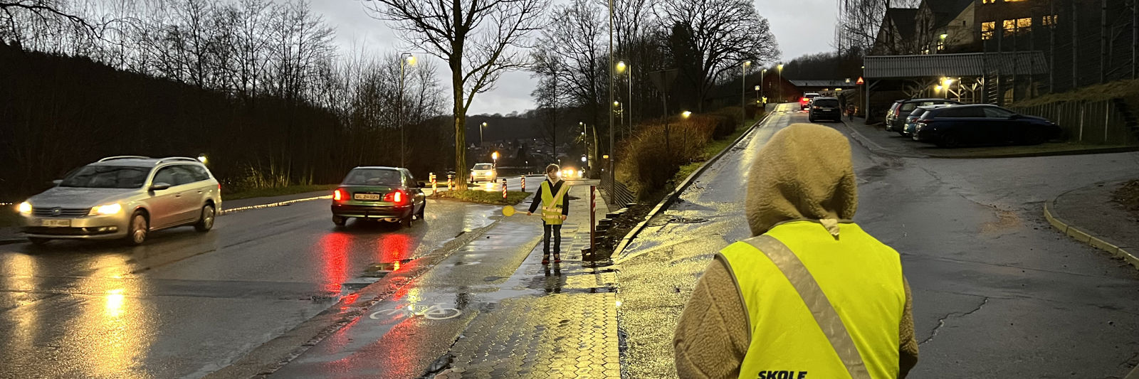 To børn står i skolepatrulje, mens biler kører forbi